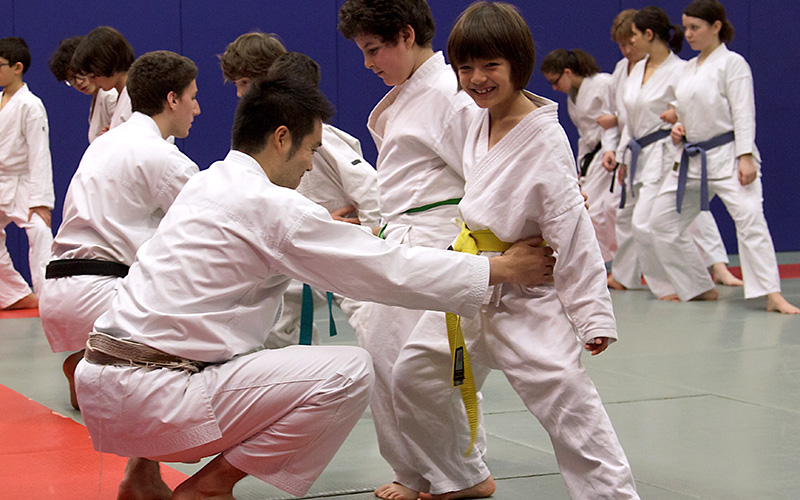 Pédagogie du karaté enfant et correction technique JKA Karaté enfant : Un instructeur corrige la position d'un jeune karatéka en ceinture jaune pendant un exercice collectif.