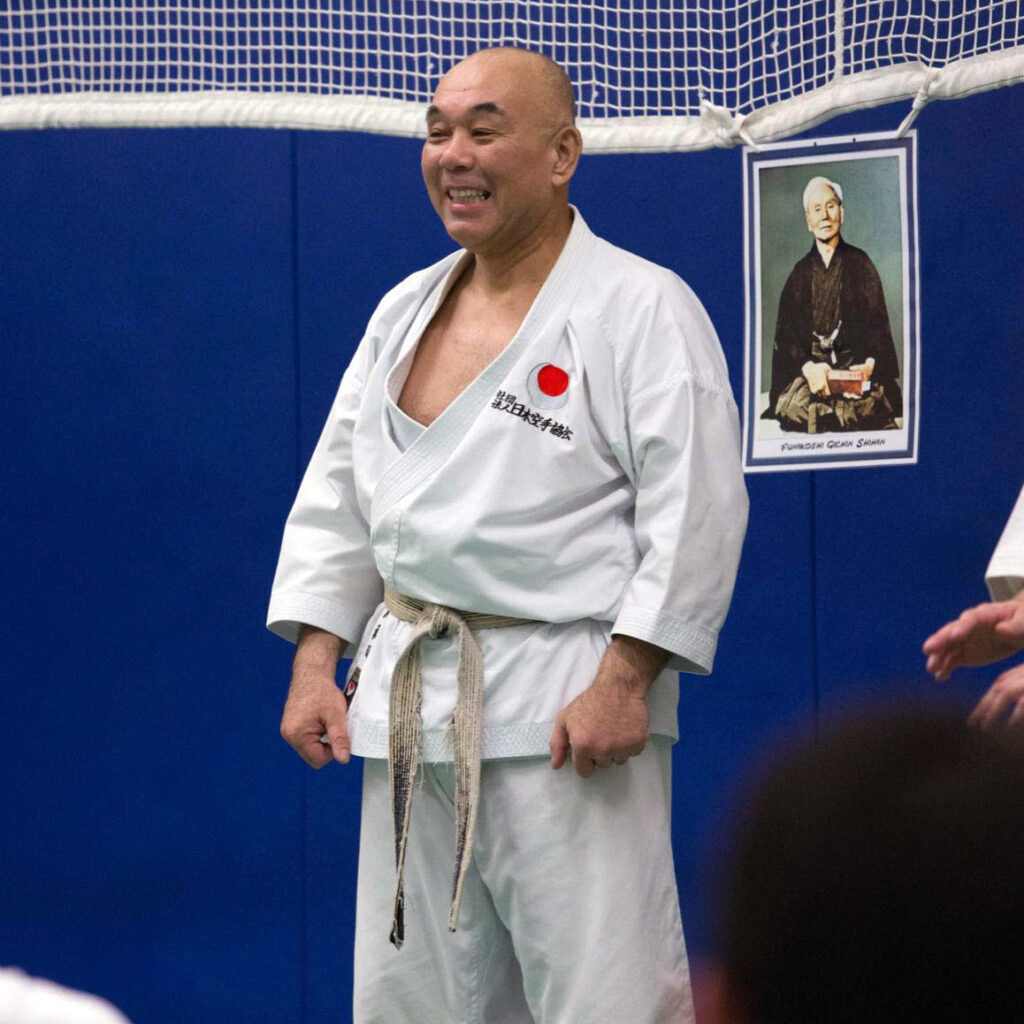 Le Maître Minoru Kawawada Sensei, grand expert JKA, souriant sur le tatami en keikogi devant un portrait traditionnel.